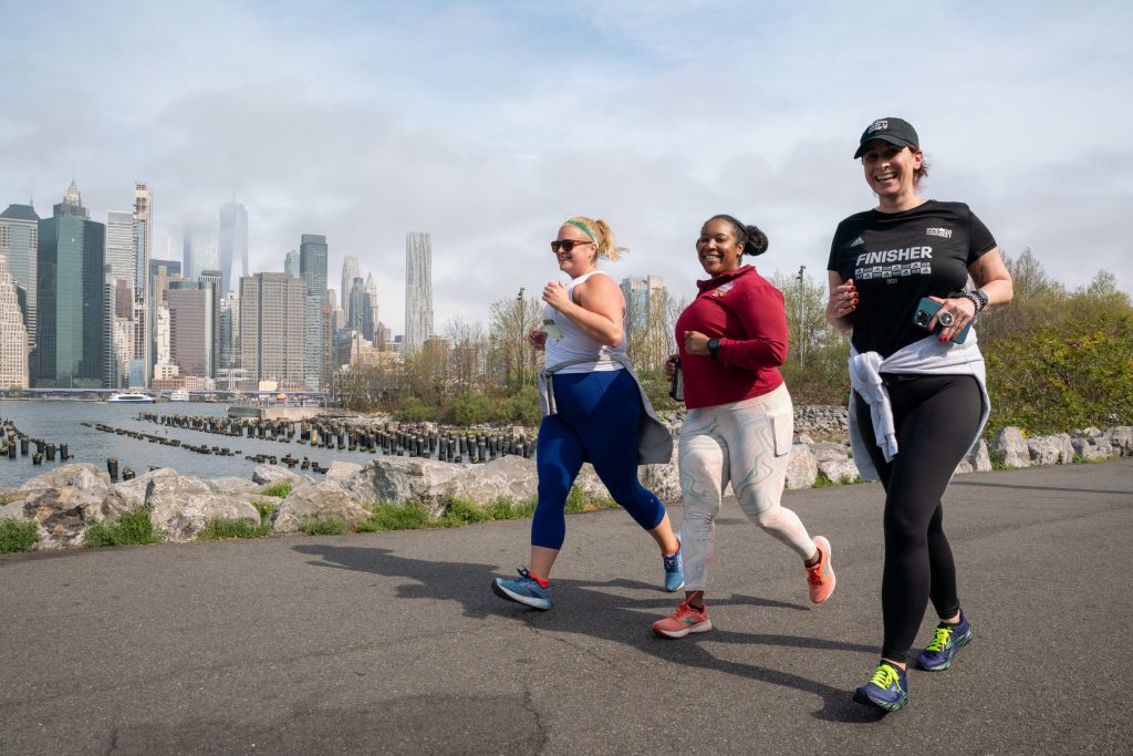 3 women slowly running along a waterside running track opposite a city in exercise gear. They're all facing the camera mid run and giving warm smiles.