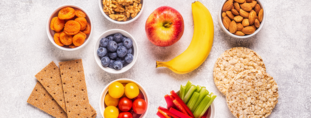 Selection of healthy snacks, including banana, apple, blueberries, nuts, crackers and vegetable sticks.