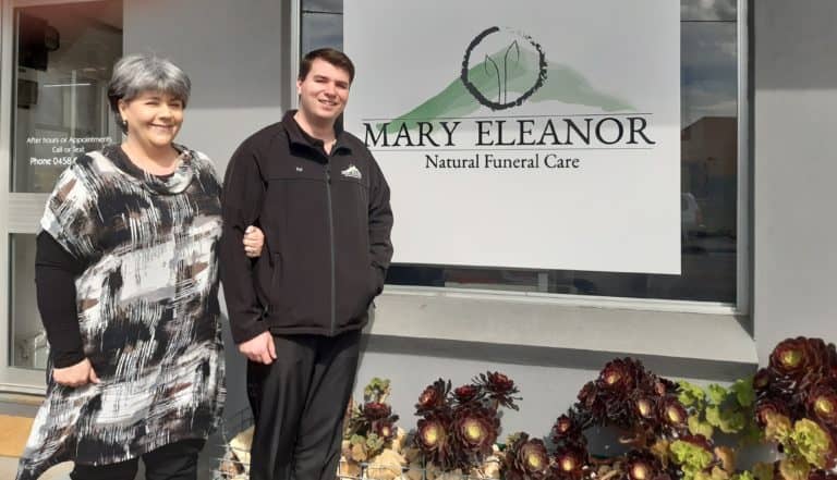 a older women and a young man stand together in front of a window and small garden. The window has a sign that says "May Eleanor". The man is wearing a zip-up back jacket and the women is wearing a black and white patterned dress.