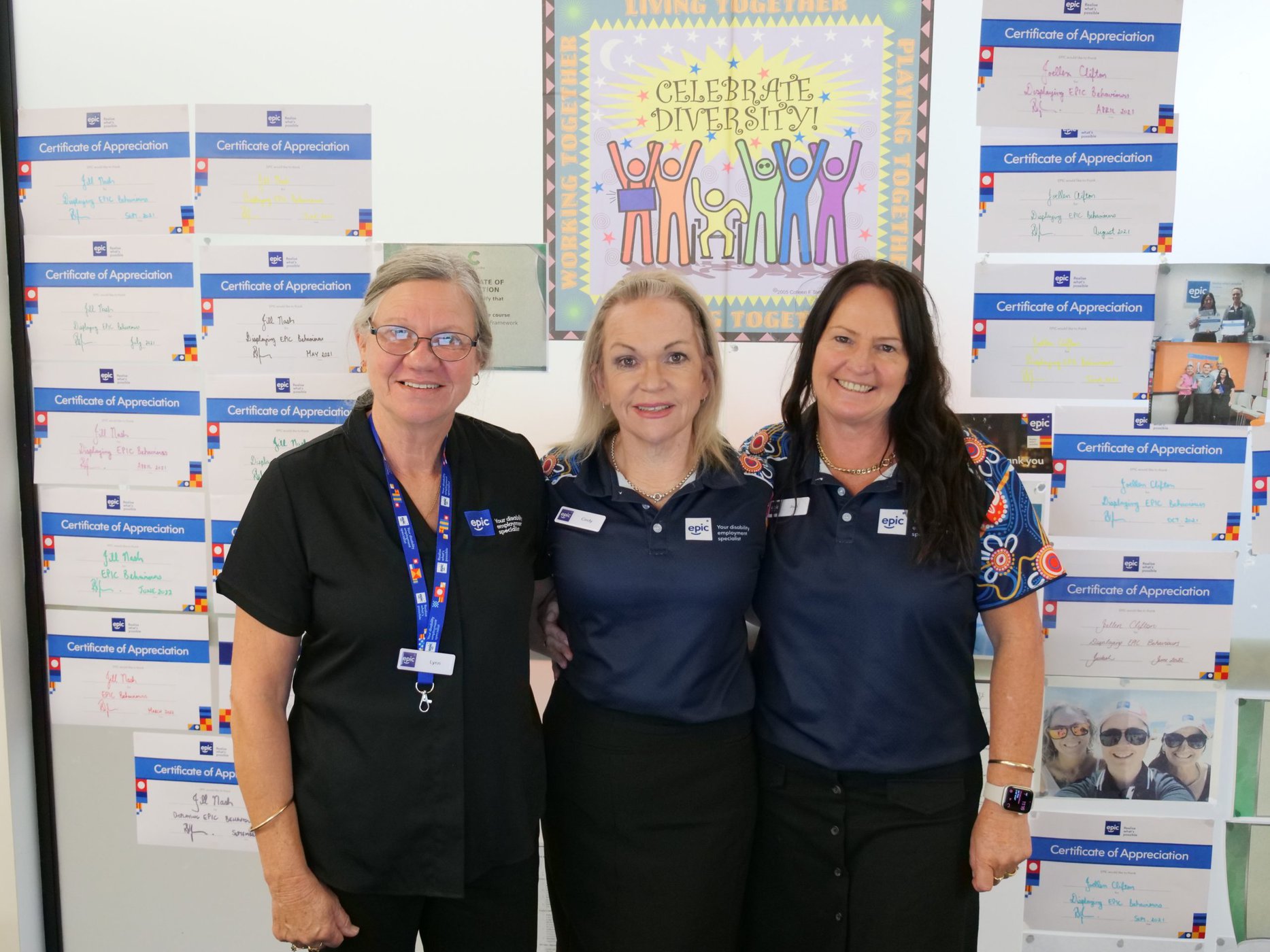Three EPIC staff members, Lyn, Cindy and Jo, smile at the camera in front of a Celebrate Diversity poster