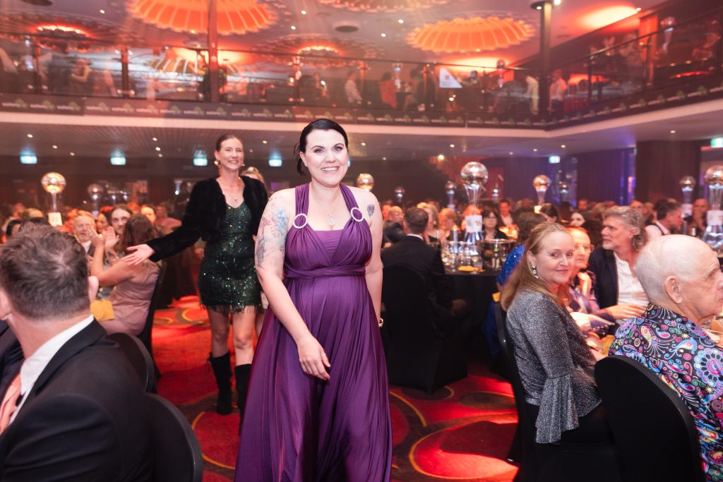 A women in a purple dress smiles at the camera as she walks through a crowd of tables at an awards ceremony