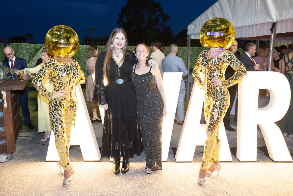 Two women dressed in black stand in between two dancers dressed in gold unitards and gold disco balls on their heads, in front of giant letters spelling out the word 'awards'.