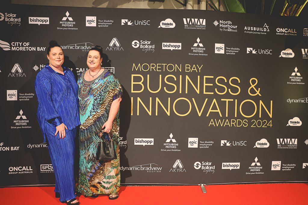 Two women stand in front of a media wall of logos at the Moreton Bay Business and Innovation awards