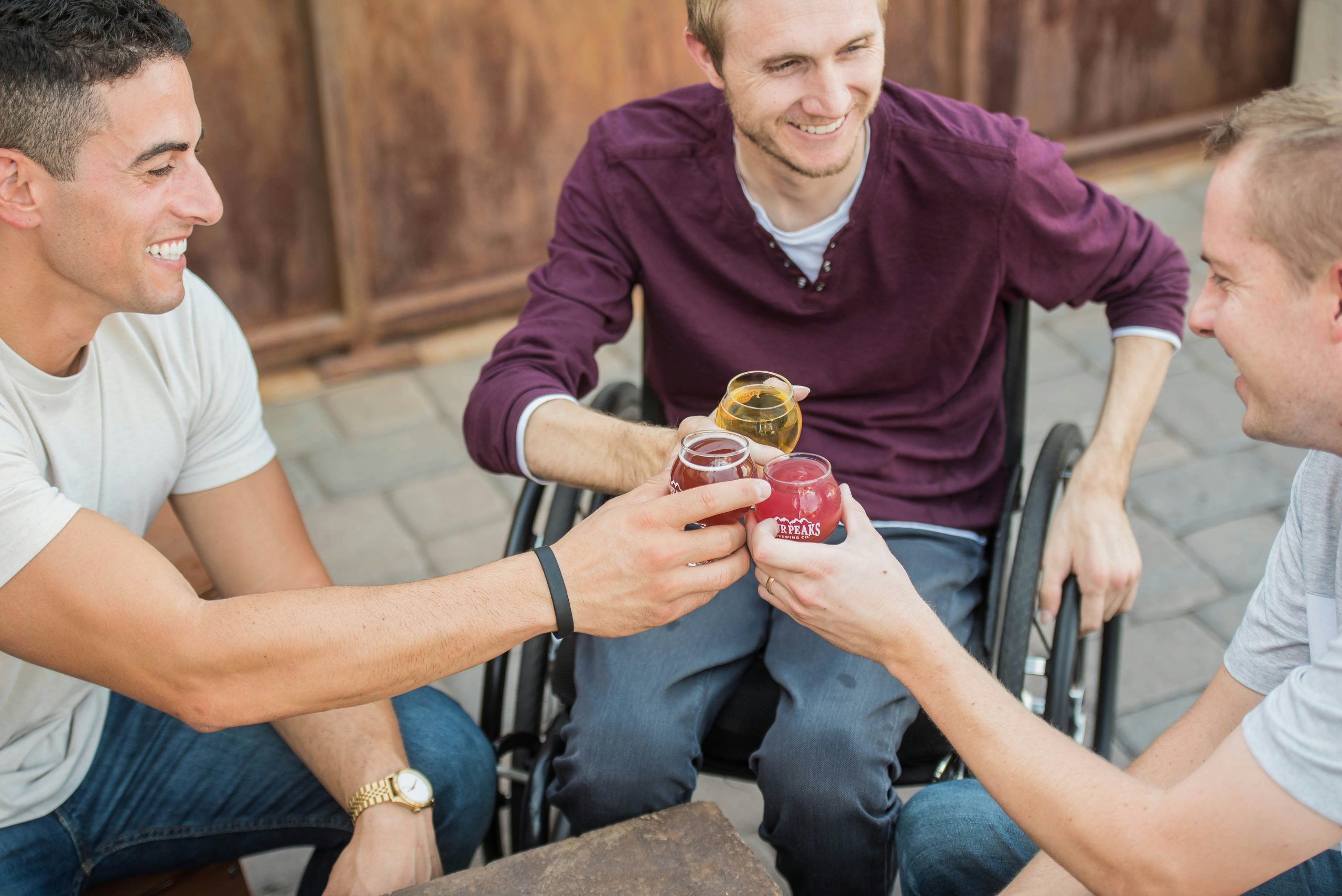 3 caucasian men cheersing their drinks. The one in the middle is sitting in a wheelchair.